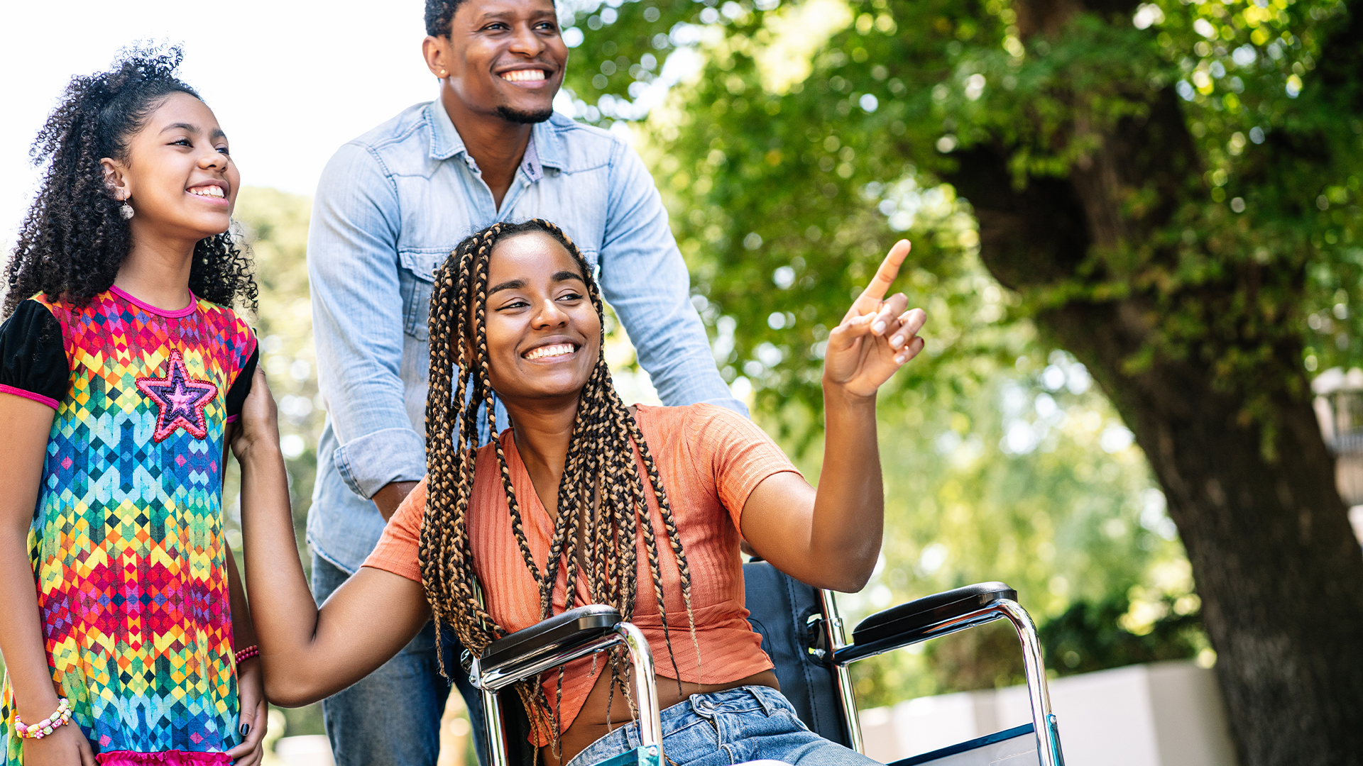A smiling woman seated in a wheelchair that is being pushed by a man. The woman is pointing out something to a young girl standing beside her.