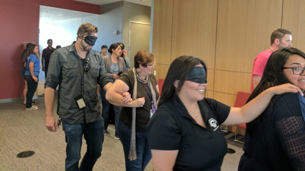 People wearing blindfolds walking side-by-side with people not wearing blindfolds