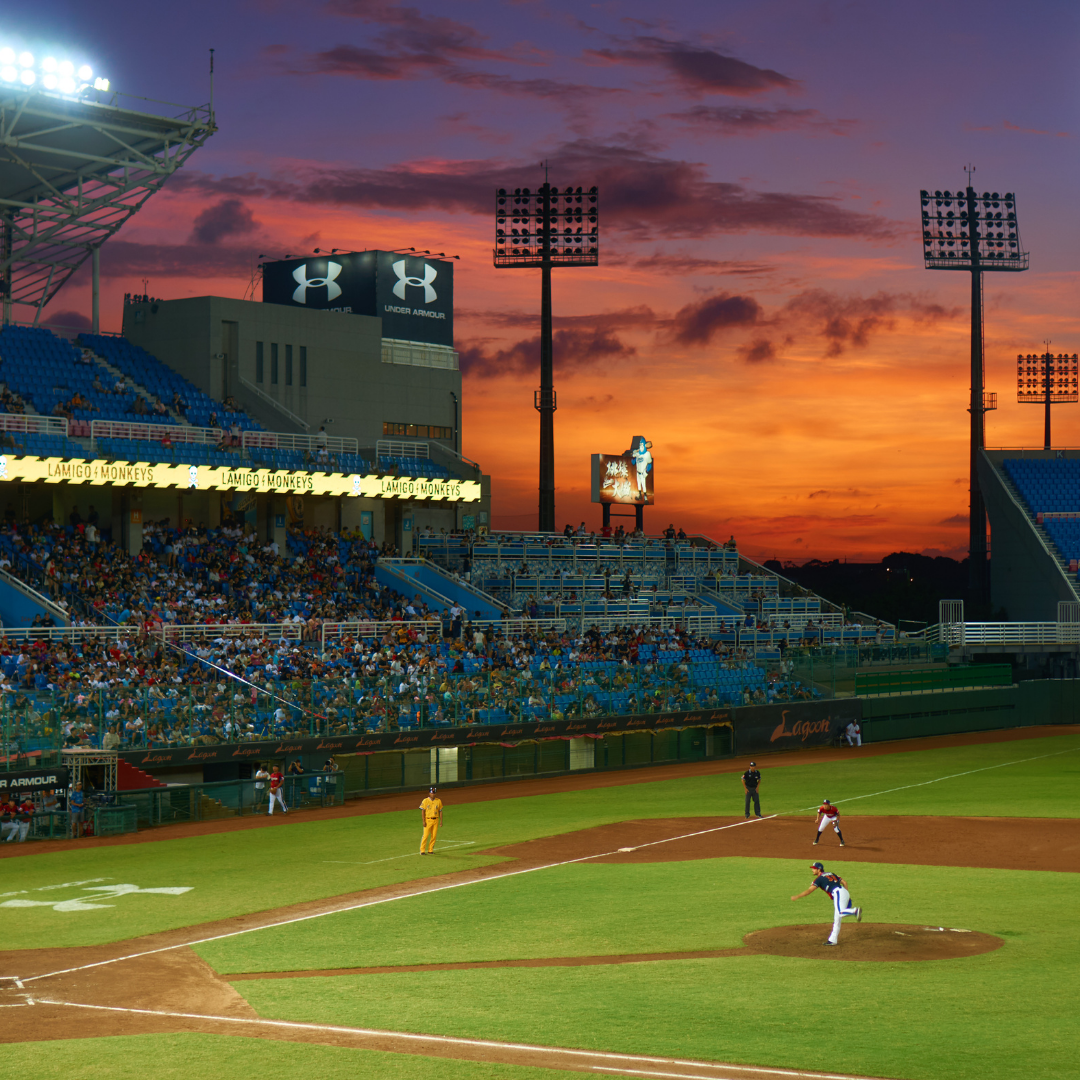A baseball game that is taking place in a mid-sized stadium. A view of the infield, one side of stadium seating, and the sky behind the stadium are shown.